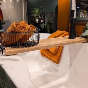 May include: A modern bathroom scene featuring a white bathtub with a wooden bath tray. Rolled orange towels are in a black wire basket and on the tray. Black candle holders and greenery add to the spa-like ambiance.
