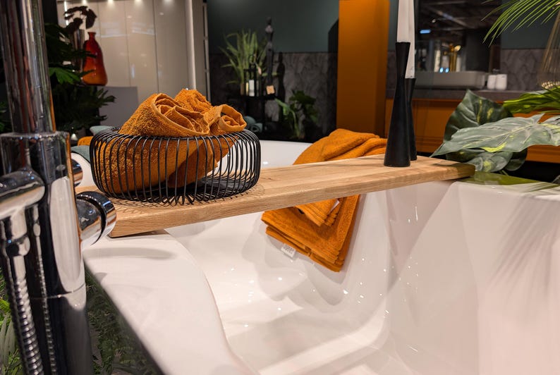 May include: A modern bathroom scene featuring a white bathtub with a wooden bath tray. Rolled orange towels are in a black wire basket and on the tray. Black candle holders and greenery add to the spa-like ambiance.