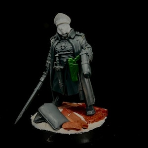 May include: A miniature figure of a soldier wearing a long dark gray coat, a peaked cap, and a chest plate with a skull emblem. The figure holds a long sword in one hand and a green pistol in a holster at the waist, standing on a base with debris.