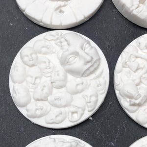 May include: White ceramic round bases with raised designs of faces. The bases are approximately 2 inches in diameter.