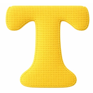 Puede incluir: Una letra "T" de ganchillo de color amarillo brillante sobre un fondo blanco. La letra mayúscula tiene una superficie texturizada y un diseño redondeado. La letra es de color sólido y parece ser un artículo decorativo.