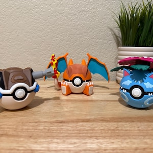 May include: Three spherical figures resembling creatures integrated into white and colored Poké Balls. The left figure is brown with gray cannons, the center is orange with blue wings and a flame tail, and the right is blue with a large pink flower.