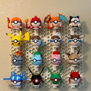 Könnte beinhalten: Eine Sammlung farbenfroher Pokémon-Charakterfiguren, die auf einer weißen, wabenförmigen Präsentation montiert sind. Die Figuren umfassen Pikachu, Glurak und andere, jeweils mit einzigartigen Designs und Farben. Die Präsentation ist an einer beigefarbenen Wand befestigt.