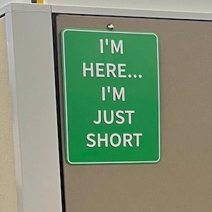 May include: A rectangular green sign with white text that reads "I'M HERE... I'M JUST SHORT." The sign is mounted on a brown wall. The sign has a white border.