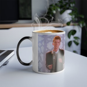 May include: A white ceramic coffee mug with a black handle. The mug features a color image of a man with red hair singing into a microphone. The background is a blurry image of a white and purple patterned design.
