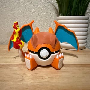 May include: A spherical orange and white creature figurine resembling a Poké Ball with blue-lined wings, small white claws, and a yellow and red flame-shaped accessory attached to its side, featuring a central black and white circular button.