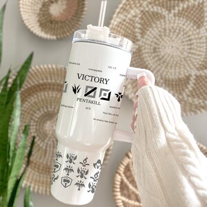 May include: White stainless steel tumbler with a black and white design featuring the words "VICTORY" and "PENTAKILL". The tumbler has a clear lid and a black straw.
