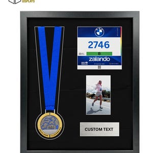 May include: A black shadow box frame containing a gold marathon medal with a blue ribbon, a race bib numbered 2746 with BMW and Zalando branding, a photograph of a runner, and a silver plaque with the text CUSTOM TEXT on a black background.