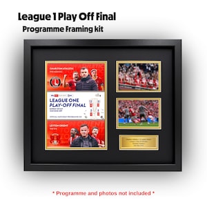 May include: Black framed kit for a League 1 Play Off Final programme. The kit includes spaces for a programme and photos. The text "League 1 Play Off Final Programme Framing kit" is at the top.