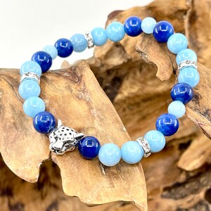 Puede incluir: Un brazalete de piedras azules y azul claro con un dije de jaguar plateado. El brazalete está hecho con cuentas redondas y tiene separadores plateados.