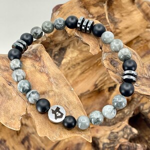 May include: A gray and black beaded bracelet with a silver charm featuring the outline of the state of Texas with a heart inside. The bracelet has black onyx beads and gray and white agate beads.