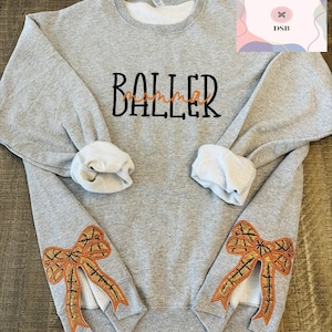 May include: Gray sweatshirt with the text "BALLER" in black embroidery. The sleeves have orange and black embroidered bows.