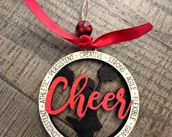 May include: A wooden ornament with a black silhouette of a cheerleader holding pom-poms. The ornament is decorated with the word "Cheer" in red and white lettering and a circle of words describing the qualities of a cheerleader, including "hard working", "creative", "strong", "agile", "flexible", "graceful", "consistent", "athletic", and "persistent".