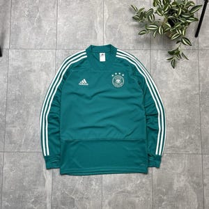 Könnte beinhalten: Türkisfarbenes Adidas-Fußballtrikot mit langen Ärmeln und weißen Streifen auf den Ärmeln. Das Trikot zeigt das Wappen der deutschen Nationalmannschaft und das Adidas-Logo. Es hat einen V-Ausschnitt.