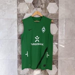 Puede incluir: Camiseta deportiva verde sin mangas con el logo de Nike y el logo del equipo Werder Bremen. La camiseta presenta el logo de Targobank en blanco, junto con detalles en blanco y una franja negra. La camiseta está colgada en una percha de madera.