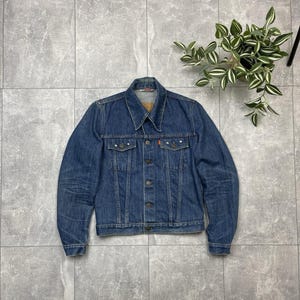 Op de afbeelding: Een klassieke blauwe spijkerjas met een knoopsluiting, twee borstzakken en een kraag. De jas heeft een versleten uiterlijk, met vervaging en subtiele slijtage. De jas is van medium wassing denim en heeft metalen klinknagels en knopen.