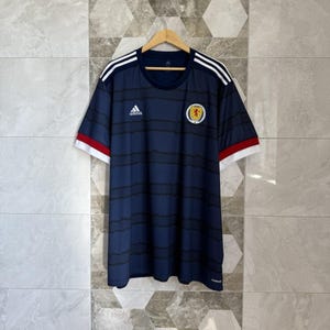 May include: Navy blue football shirt with black horizontal stripes. The shirt has white shoulder stripes, a round neck, and red and white sleeve trim. The Adidas logo is on the chest, and a Scottish emblem is on the left side.