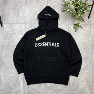 Op de afbeelding: Zwarte hoodie met het woord "ESSENTIALS" in witte blokletters over de borst. De hoodie heeft een kangoeroezak en een klein label aan de capuchon. Het kledingstuk is gemaakt van gebreid materiaal.