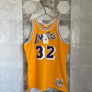 Puede incluir: Una camiseta de baloncesto amarilla con ribetes morados y blancos. El número "32" está en la parte delantera de la camiseta en morado y blanco. La palabra "Lakers" está en la parte delantera de la camiseta en morado. La camiseta tiene una etiqueta que dice "NBA Classics" y "Mitchell & Ness".