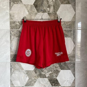May include: Bright red athletic shorts with an elastic waistband, featuring a team logo on the left leg and the Reebok logo on the right. The shorts are hanging on a metal hanger against a patterned tile background.