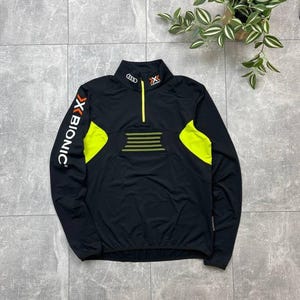 Puede incluir: Chaqueta deportiva negra con detalles en amarillo neón. Cuello alto con cremallera amarilla. El texto "X BIONIC" en blanco en la manga izquierda. Diseño deportivo.