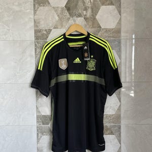 May include: Black soccer jersey with neon yellow stripes on the shoulders and chest. The jersey features the Spanish national team crest, Adidas logo, and FIFA emblem. The shirt is hanging on a wooden hanger.