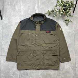 May include: Olive green and black outdoor jacket with multiple pockets and a high collar. The jacket features a button-up front and a small logo patch on the chest. The jacket is made of a durable, weather-resistant material.