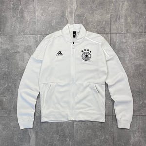 Könnte beinhalten: Weiße Jacke mit Reißverschluss, Adidas-Logo und dem Emblem der deutschen Nationalmannschaft. Die Jacke hat einen Stehkragen, zwei Vordertaschen und einen durchgehenden Reißverschluss. Das Material ist weich.