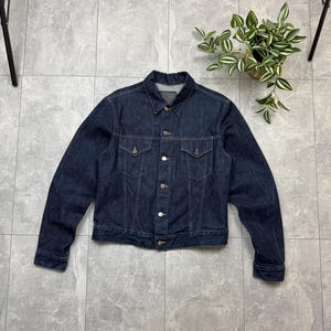 May include: A dark blue denim jacket with a button-up front, two chest pockets, and two side pockets. The jacket has a classic collar and long sleeves. The denim is a solid, dark wash. The jacket is laid flat on a gray tiled surface.