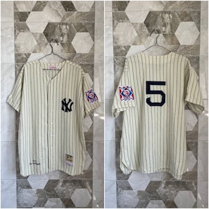 Puede incluir: Camiseta de béisbol color crema con finas rayas azul marino. Presenta el logo de los Yankees de Nueva York en el pecho y el número 5 en la espalda. Incluye parches en las mangas y una etiqueta en la parte inferior.