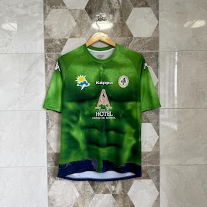 May include: A green short-sleeve jersey with the Kappa logo and the text "A HOTEL CIUDAD DE ASTORGA". The jersey has a design that resembles a muscular torso. A sun and cloud graphic is on the left chest.