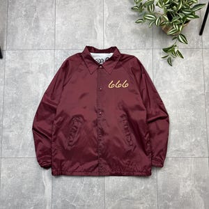 May include: A maroon coach jacket with a button-up front and a collar. The number "666" is embroidered in a gold colour on the front. The jacket is made of a smooth material, possibly nylon.