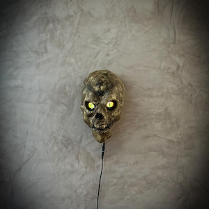 Peut inclure: Un crâne doré avec des yeux jaunes brillants est suspendu contre un mur gris texturé. Le crâne a une apparence vieillie, avec des détails visibles dans la structure osseuse. Un cordon noir s'étend de la base du crâne.