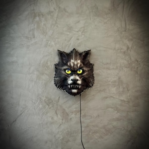Peut inclure: Décoration murale en forme de tête de loup noire et grise, avec des yeux jaunes brillants et des dents blanches acérées. La tête de loup est fixée sur un mur gris, avec un cordon d'alimentation noir qui descend. Aspect texturé et réaliste.