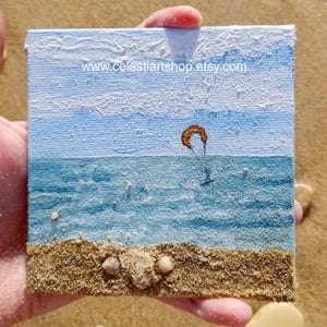 Puede incluir: Arte en lienzo cuadrado que representa una escena de playa con un kitesurfista. El cielo es azul claro con nubes blancas texturizadas. El océano es azul texturizado y la playa es arenosa con pequeñas conchas. El sitio web www.celestiartshop.etsy.com está impreso.