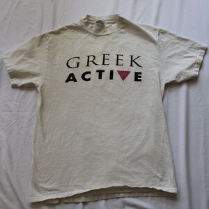 May include: A short-sleeved, off-white crew neck t-shirt featuring the text GREEK ACTIVE printed in black across the chest. The letter V in the word ACTIVE is replaced by an inverted pink triangle. The shirt has a standard fit and hem.