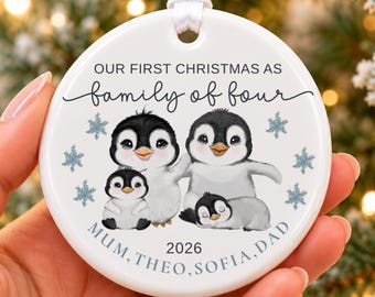 May include: A round white ceramic ornament with a white ribbon hanger. The design features four illustrated penguins, blue snowflakes, and the text: OUR FIRST CHRISTMAS AS family of four, MUM, THEO, SOFIA, DAD, and 2026.