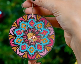 May include: A round Christmas ornament with a colorful floral pattern and a small bird in the center. The ornament is held by a hand against a green Christmas tree background.