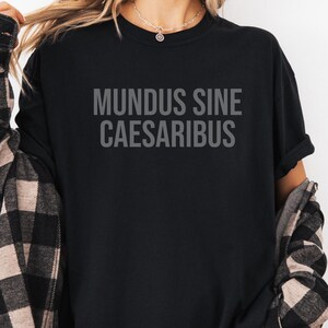 May include: A black t-shirt with the text "MUNDUS SINE CAESARIBUS" printed in white on the front. The shirt is being worn by a person with long brown hair and a plaid shirt tied around their waist.
