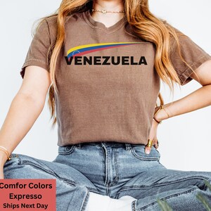 May include: A brown t-shirt with a yellow, blue, and red graphic of the Venezuelan flag and the word "VENEZUELA" in black text.