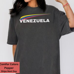 May include: A dark gray t-shirt with a yellow, blue, and red arc design and the word "VENEZUELA" in white text.