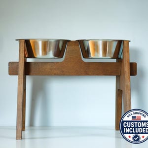Può includere: Un alimentatore per animali domestici in legno con due ciotole in acciaio inossidabile. L'alimentatore è realizzato in legno marrone scuro e ha un design semplice e robusto. Le ciotole sono inserite nel telaio di legno.