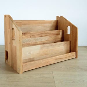 May include: A light-colored wooden bookshelf with three shelves. The bookshelf has a rectangular shape with angled sides and cut-out handles. The wood grain is visible, and the shelves are at different levels.