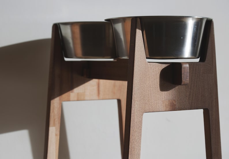 May include: A wooden pet feeder with two stainless steel bowls. The feeder has a light brown, natural wood finish and a minimalist design. The bowls are elevated, providing a comfortable feeding height for pets. The feeder is designed for small to medium-sized pets.