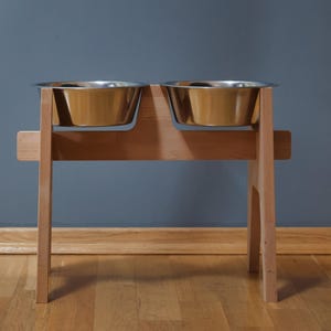 May include: A wooden pet feeder with two stainless steel bowls. The bowls are set into a light-colored wooden frame with a simple, modern design. The feeder is elevated, with a sturdy base and legs.