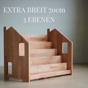 May include: A light-colored wooden bookshelf with a house-shaped design. The bookshelf has five shelves and features cut-out windows. The text "EXTRA BREIT 70cm 5 EBENEN" is displayed at the top of the image.