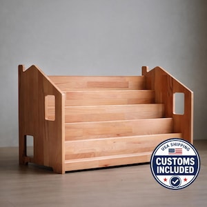 Op de afbeelding: Een houten boekenplank met een trapontwerp. De boekenplank heeft vier treden en is gemaakt van lichtgekleurd hout. Het heeft uitgesneden handgrepen aan de zijkanten en een huisvormige bovenkant. De boekenplank is ontworpen voor kinderboeken.