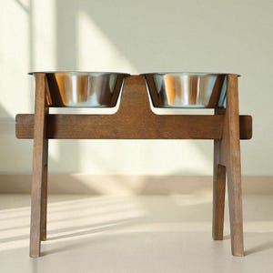 May include: A wooden pet feeder with two stainless steel bowls. The feeder is made of dark brown wood and has a simple, modern design. The bowls are elevated, making it easier for pets to eat and drink. The feeder is suitable for cats and dogs.