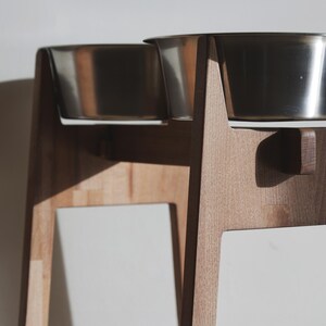 May include: A wooden pet feeder with two stainless steel bowls. The feeder has a natural wood finish and a minimalist design. The bowls are elevated, providing a comfortable eating height for pets. The feeder is designed for small to medium-sized dogs or cats.