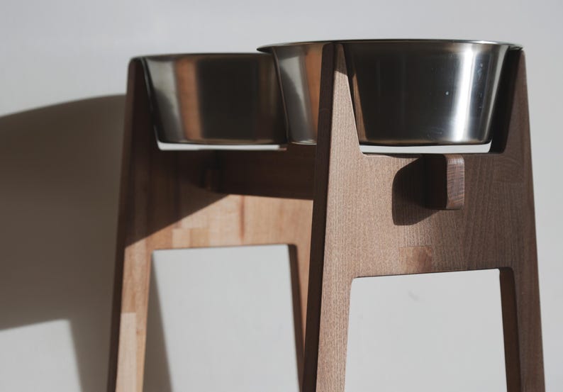 May include: A wooden pet feeder with two stainless steel bowls. The feeder has a natural wood finish and a minimalist design. The bowls are elevated, providing a comfortable eating height for pets. The feeder is designed for small to medium-sized dogs or cats.